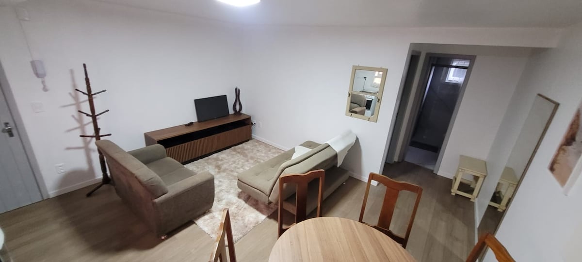 An open living area is displayed, featuring a light-colored rug under a modern sectional sofa and a wooden media console with a television. A dining table with wooden chairs is visible, alongside a floor mirror that reflects the bright, neutral tones of the space.