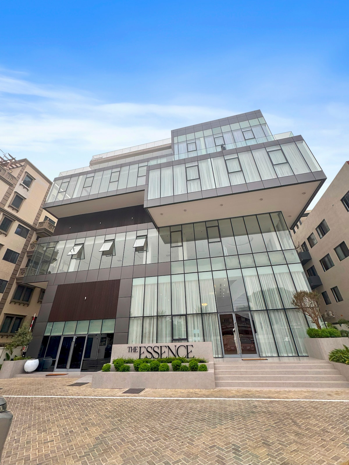 An elegantly designed building features multiple levels with expansive glass windows, allowing natural light to flood the interiors. The modern façade is complemented by neatly trimmed landscaping at the entrance, highlighting the stylish atmosphere of the Essence Apartments.