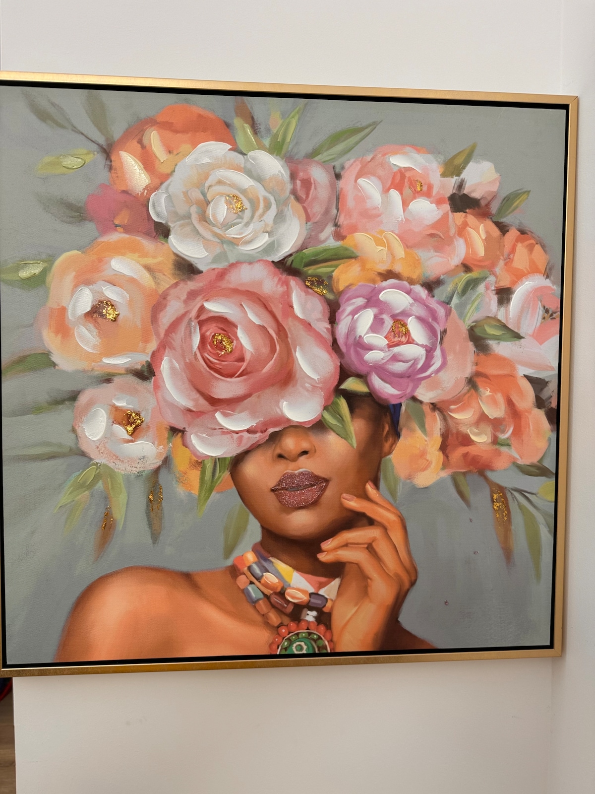 A framed artwork features a figure adorned with a vibrant floral headdress, showcasing large blossoms in shades of pink, peach, and white. The background is a soft grey, providing contrast to the rich colors of the flowers. A decorative necklace adds an additional element of interest.