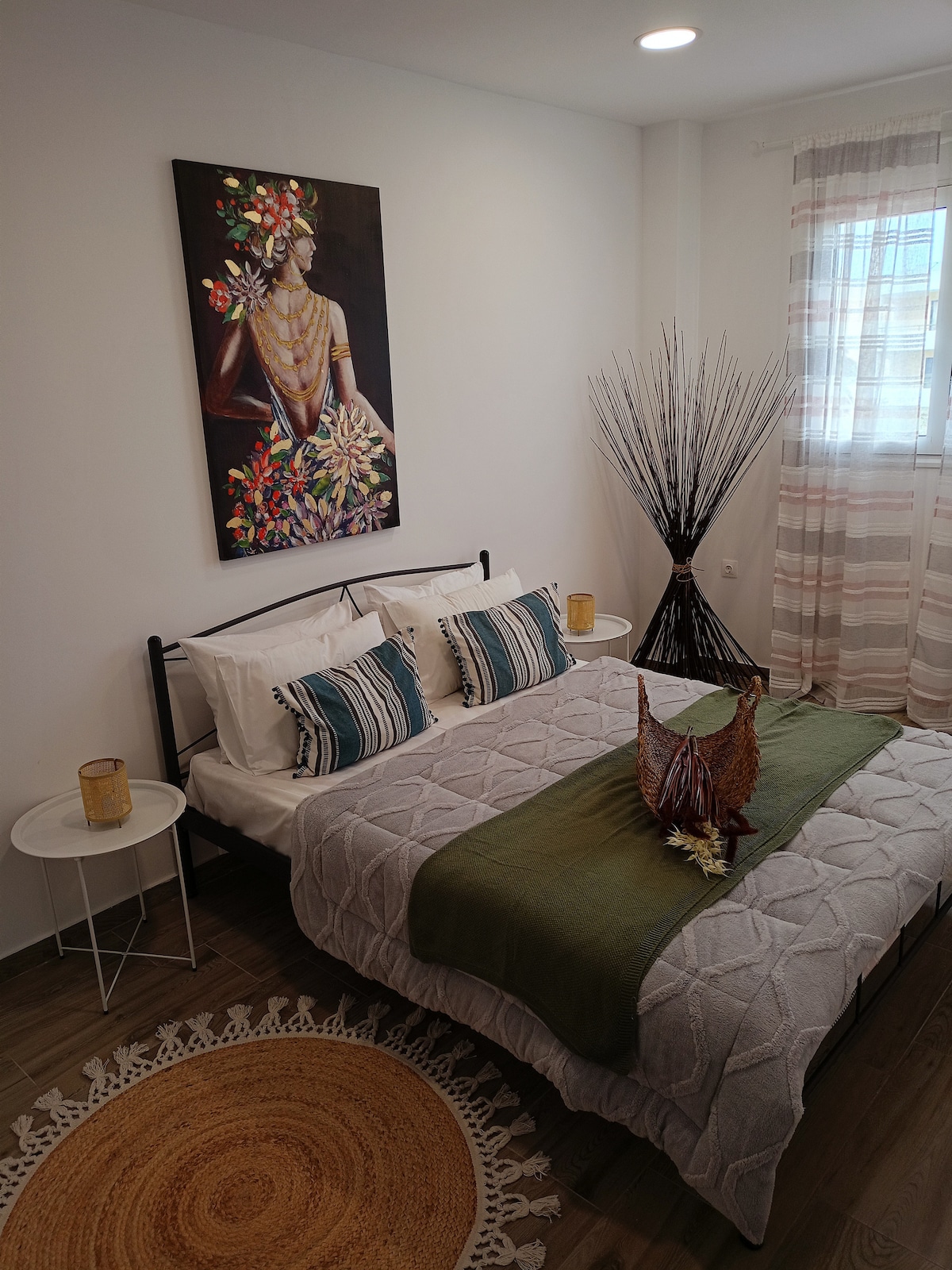 A comfortable sleeping area showcases a spacious bed adorned with textured linens and accent pillows. A wall art piece adds a splash of color, while a large decorative vase and a round rug complement the room's design. Natural light filters through sheer curtains.