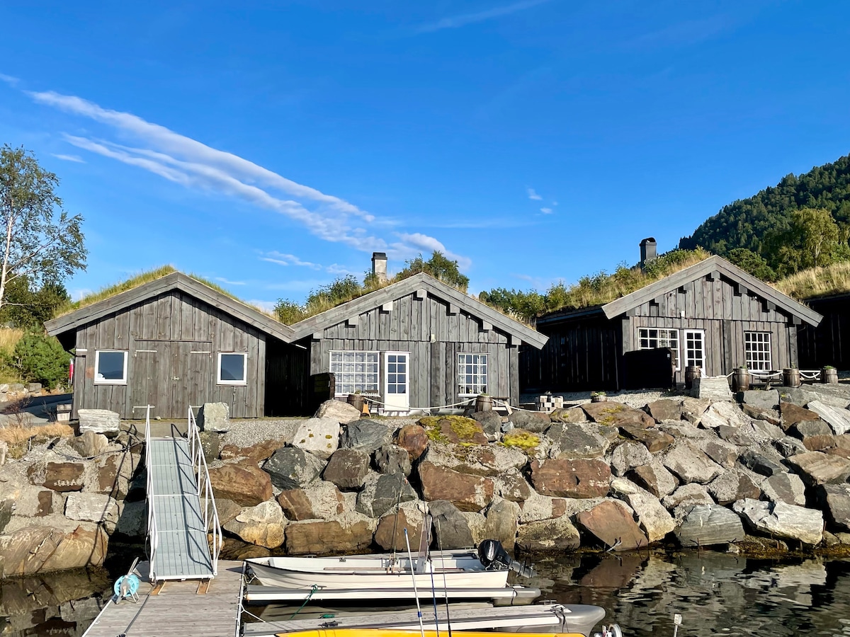 A cluster of rustic wooden cabins is set against a clear blue sky, with a stony shoreline in the foreground. Each cabin features large windows and grass-roofed designs, blending harmoniously with the natural surroundings. Small boats are visible docked along the water's edge.