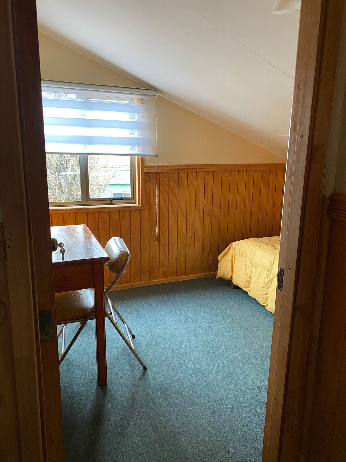 A compact room features wooden paneling and blue carpeting. A spacious bed with a yellow coverlet is positioned against one wall, and a small desk with a chair is situated near the window, allowing natural light to illuminate the space.