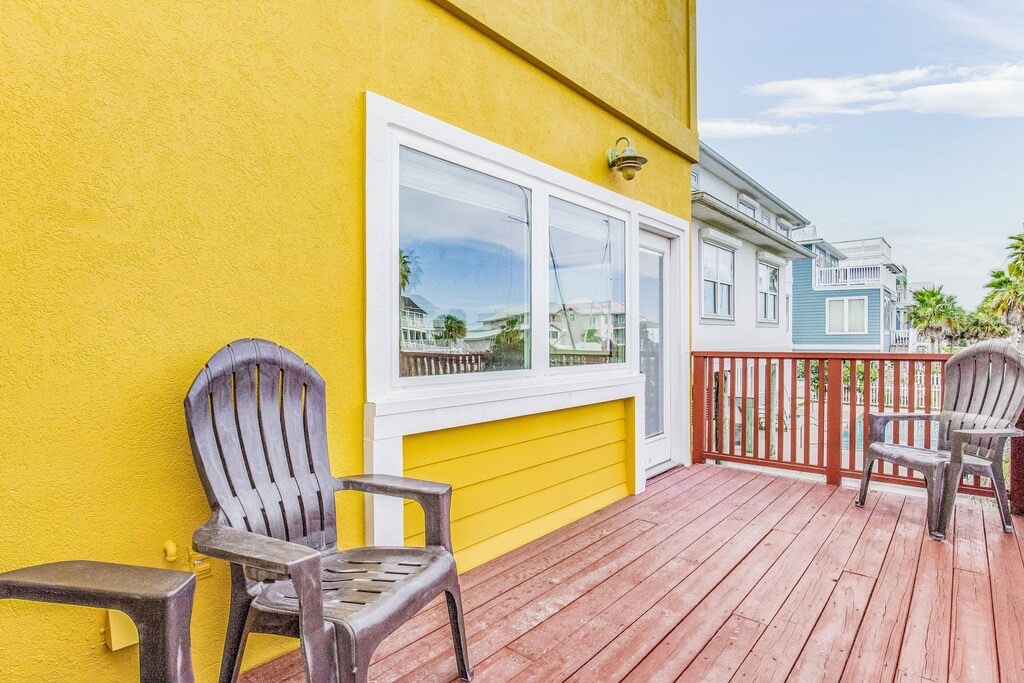 A deck with wooden flooring features two black chairs facing the vibrant yellow wall of the building. A large window provides natural light and views of the surroundings, enhancing the outdoor space for relaxation.