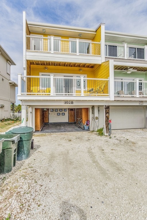 A three-story house painted in bright yellow is shown, featuring a covered balcony on the second level. The ground level includes a garage with open doors revealing a tidy space, while a gravel driveway leads up to the property, flanked by green waste bins.