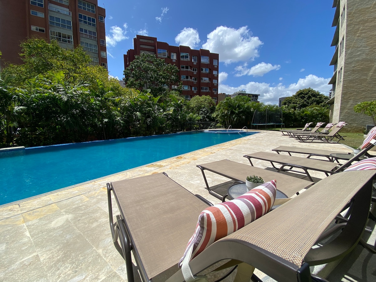 A large outdoor swimming pool is bordered by lounge chairs, arranged on a stone patio. Lush greenery surrounds the pool area, offering a tranquil environment. Clear blue skies and scattered clouds create a bright and inviting atmosphere.