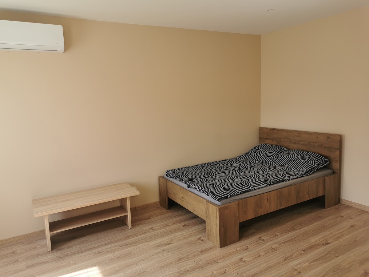 A spacious sleeping area features a wooden bed frame with a patterned mattress. A simple wooden bench is positioned nearby on light-colored flooring, complemented by neutral walls and ample natural light.