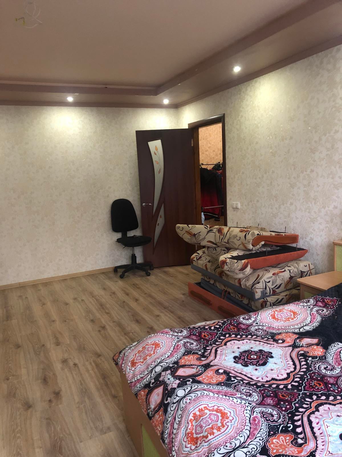 A spacious room features a patterned bedspread on a sizable bed, complemented by a decorative couch with floral upholstery. A black chair is positioned against a wall. Soft lighting emanates from recessed ceiling fixtures, illuminating the light-colored walls and wooden floor.