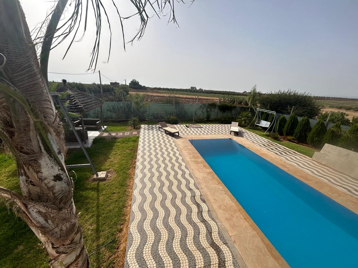 A serene outdoor area features a clear swimming pool surrounded by well-maintained lawn and patterned tiles. Palm trees provide shade, while loungers are positioned nearby for relaxation. The landscape offers unobstructed views of the surrounding greenery and distant fields.