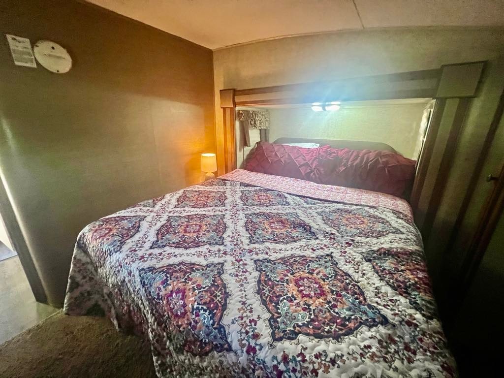 A comfortable bedroom features a queen-sized bed adorned with a patterned quilt. Soft lighting from bedside lamps creates a gentle glow, while a small clock is mounted on the wall. The warm tones of the walls contribute to a cozy ambiance.