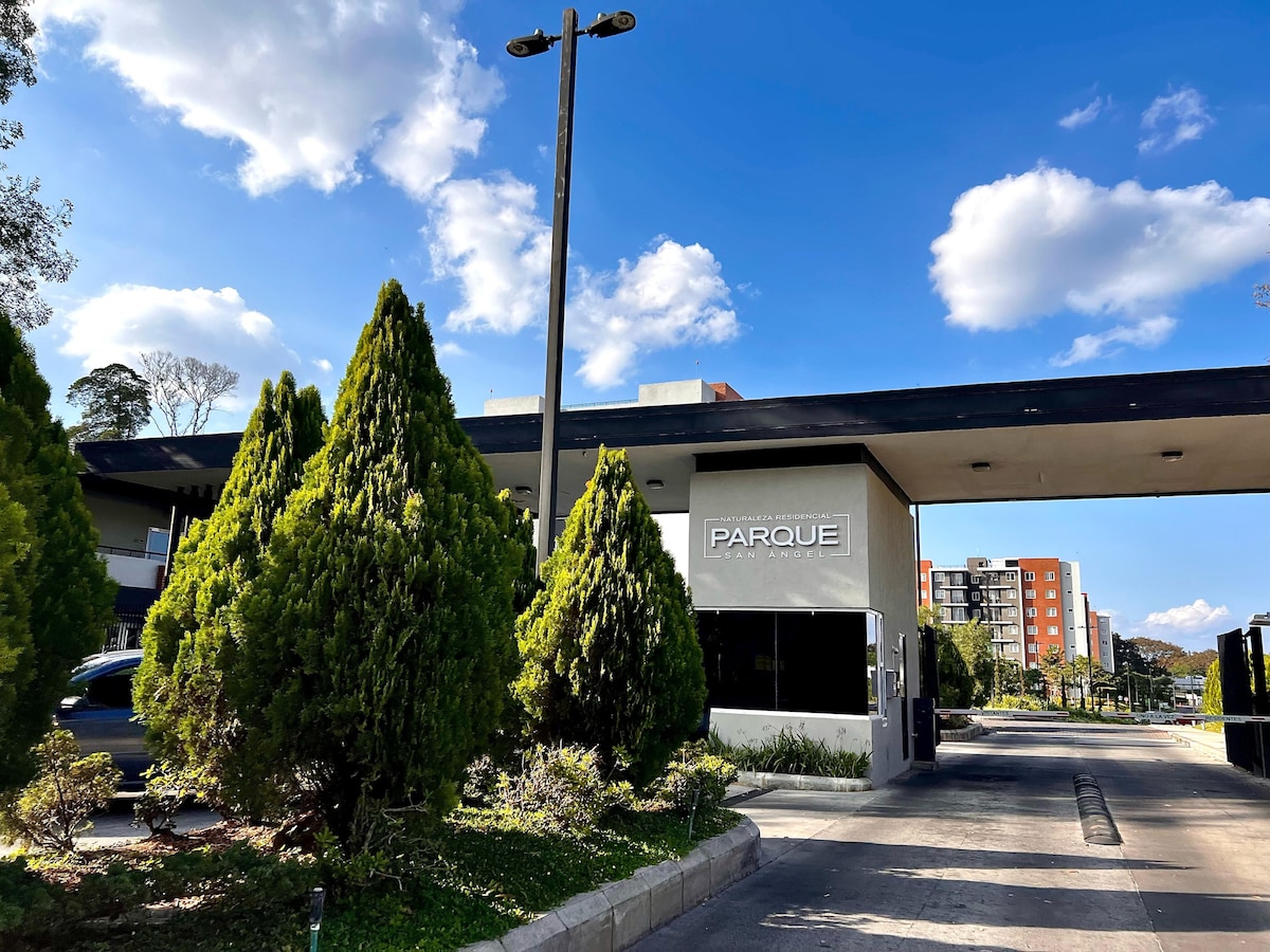 An entrance to a gated community is depicted, featuring well-maintained greenery including tall conical trees. A modern sign labeled 'PARQUE' is prominently placed on the building structure. The background reveals low-rise buildings under a clear blue sky dotted with a few clouds.