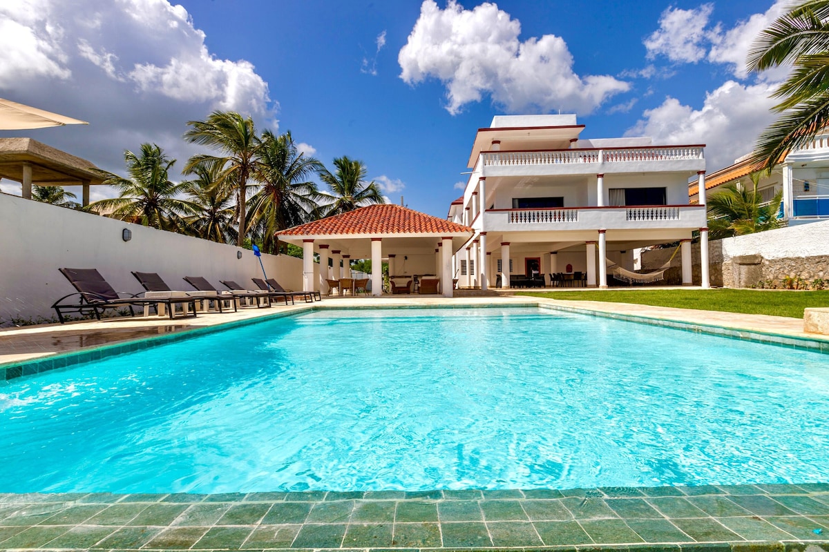 A spacious outdoor area features a clear blue swimming pool surrounded by lounge chairs. In the background, a multi-level white building with a red-tiled roof is visible, framed by palm trees and complemented by a vibrant blue sky with scattered clouds.