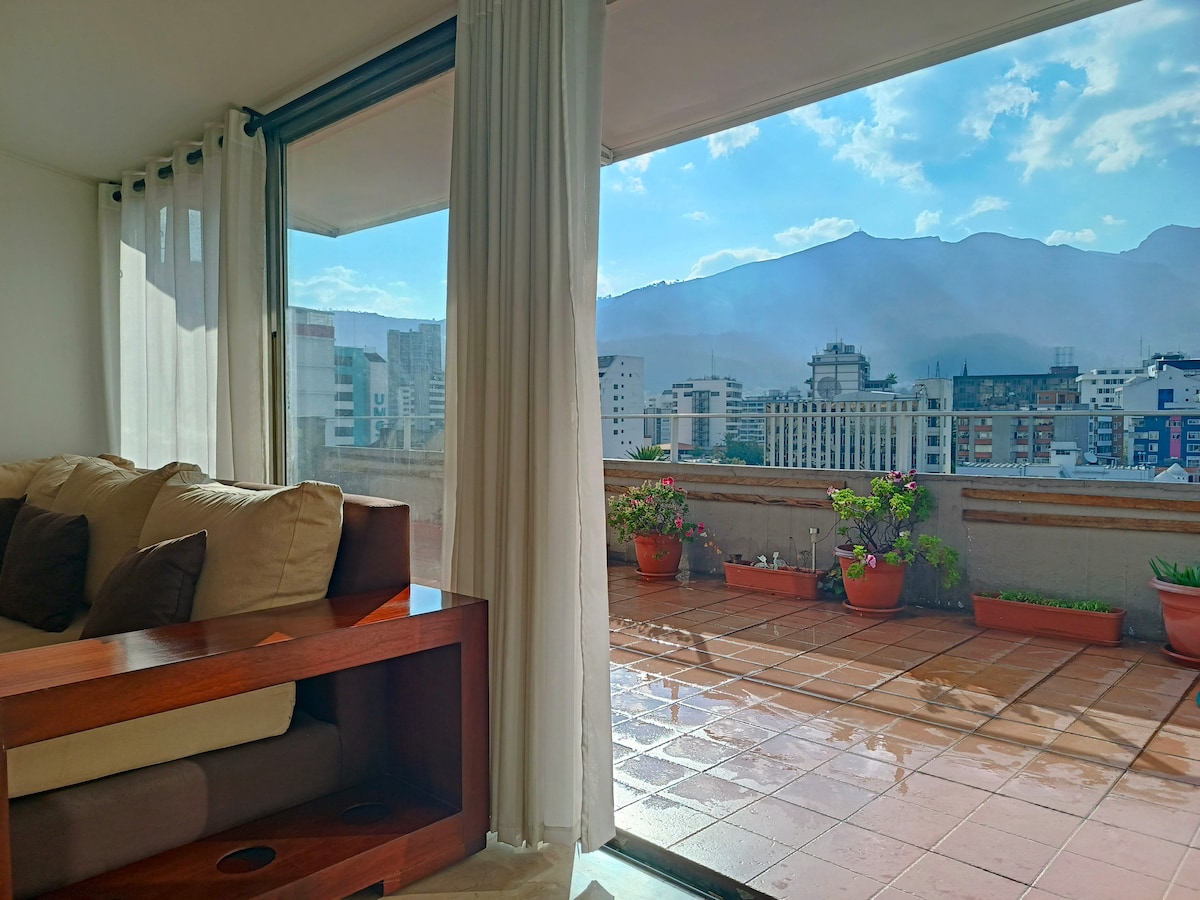 A cozy living area features a large window with sheer curtains, allowing natural light to flow in. Outside, a spacious terrace is visible, adorned with potted plants and offering expansive views of the city and mountains under a bright sky.