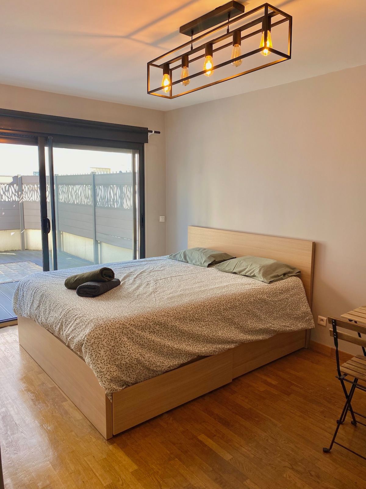A bedroom with a large bed featuring a light-colored duvet and two pillows. Natural light enters through sliding glass doors that lead to a terrace. A compact dining table and two chairs are positioned nearby, enhancing the room's functionality.
