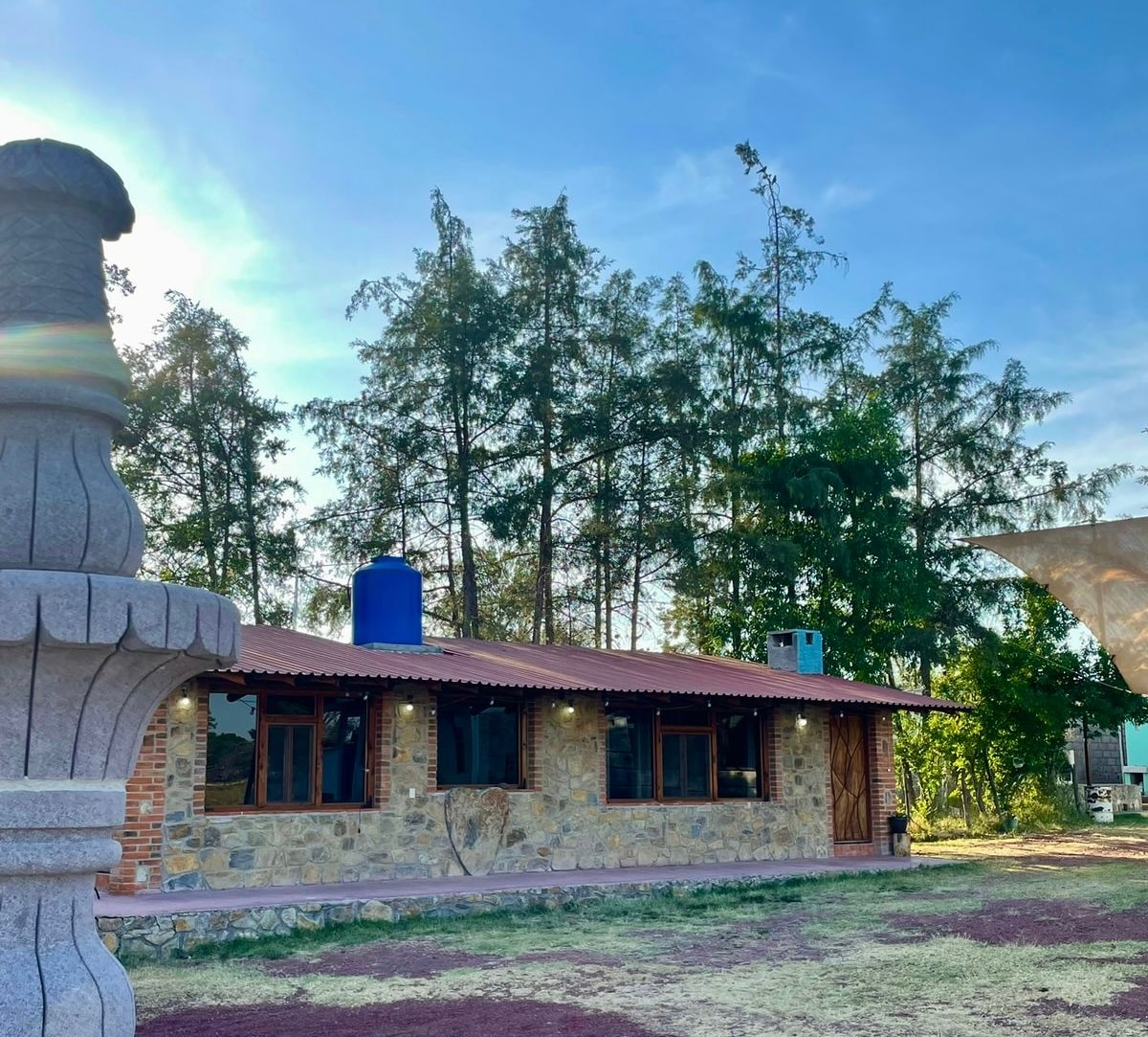 A rustic stone building is situated amidst tall trees, featuring a red-tiled roof and multiple large windows that allow natural light to fill the interior. A water tank is visible on the roof, enhancing the building's functionality.