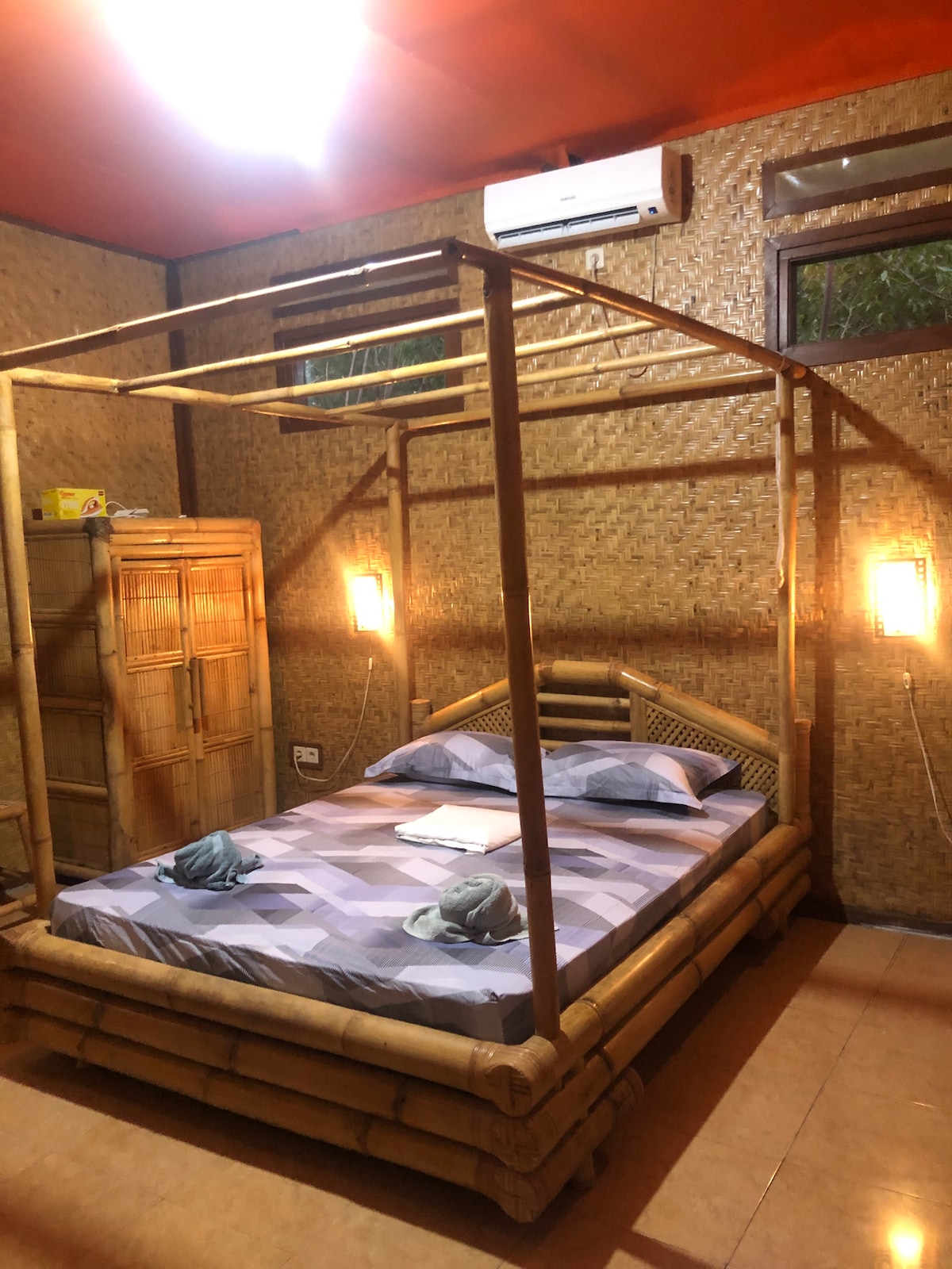 A bamboo four-poster bed is centered in a room with textured walls and natural elements. Soft lighting is provided by wall sconces on either side. Windows allow some natural light, while a wooden wardrobe is positioned nearby.