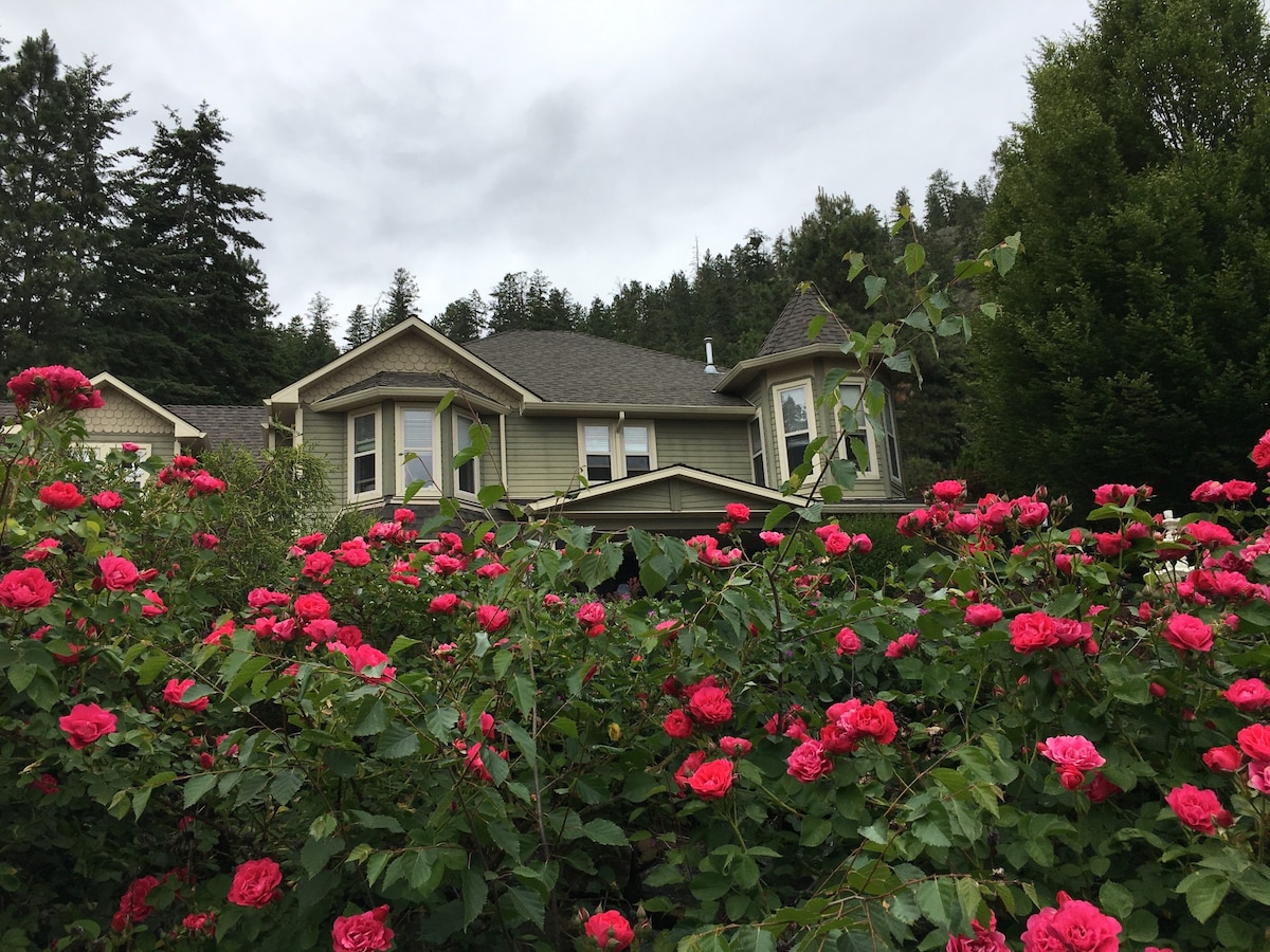 The exterior of a charming house is framed by vibrant pink roses, creating a lush garden setting. The architecture features multiple gables and large windows, showcasing the inviting façade against a backdrop of green trees and a cloudy sky.