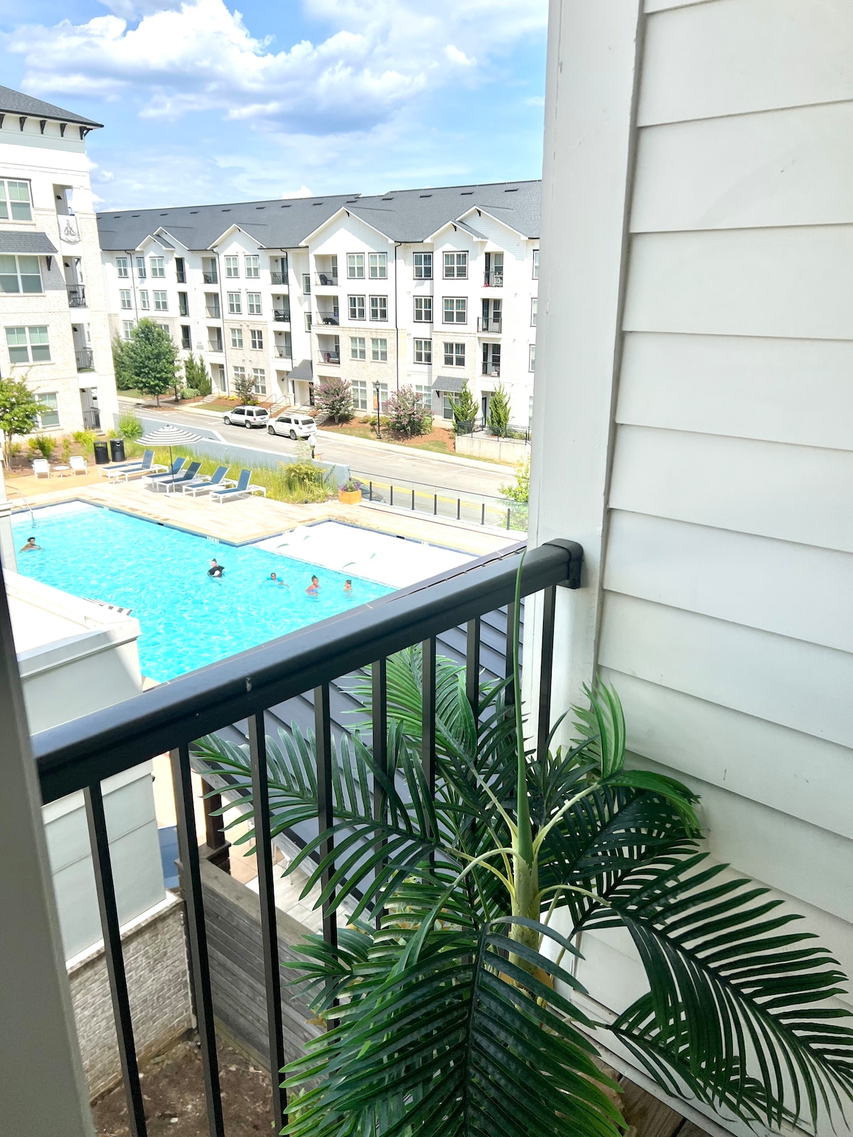 A view of the community's saltwater pool is visible from the balcony, surrounded by lounge chairs and greenery. The pool area is framed by modern apartment buildings, with a clear blue sky and soft clouds overhead.