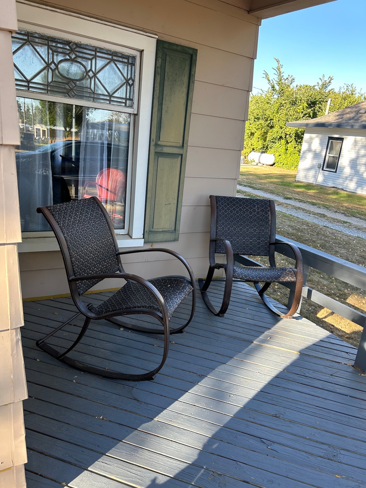 A porch area is shown, featuring two rocking chairs on a wooden deck. Natural light filters through a nearby window with decorative glass. Surrounding greenery is visible, enhancing the outdoor space.