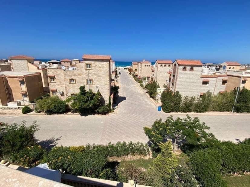 A view from an elevated position showcases a paved road lined with well-maintained stone buildings and greenery. In the distance, the blue sea is visible, complementing the tranquil surroundings. The area offers a sense of peace and accessibility to the beach.