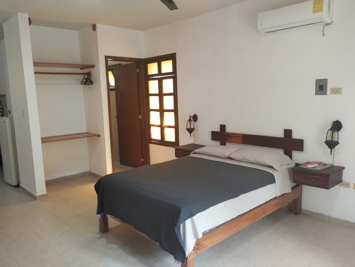A minimalist sleeping area features a wooden bed with a gray blanket and simple bedside tables. Ambient light filters through a window with white panes. Additional shelving is visible on one wall, and a door leads to a small hallway.