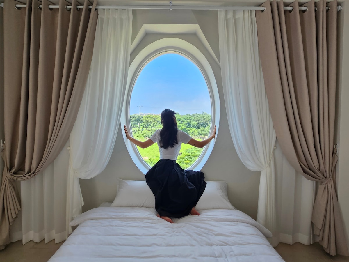 A spacious bed is set against a large, circular window that frames a scenic view of surrounding greenery. Light drapes in soft colors adorn the window, while a person sits gracefully on the bed, hands placed on the window ledge, gazing outside.