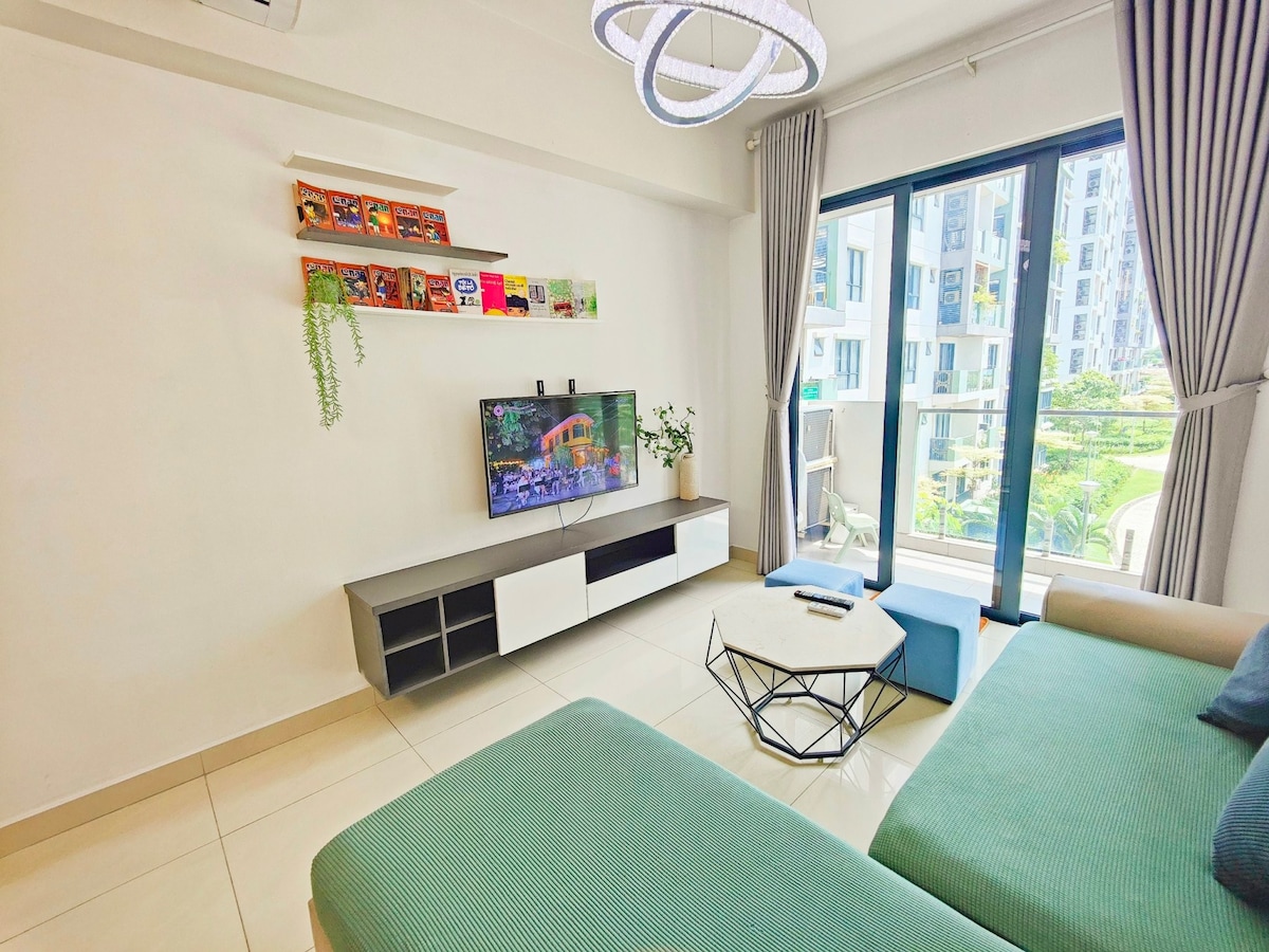 A bright living area features a green sectional sofa and a modern coffee table. A television is positioned on a sleek unit with storage, while a shelf holds an array of books. Large glass doors open to a view of the garden outside.
