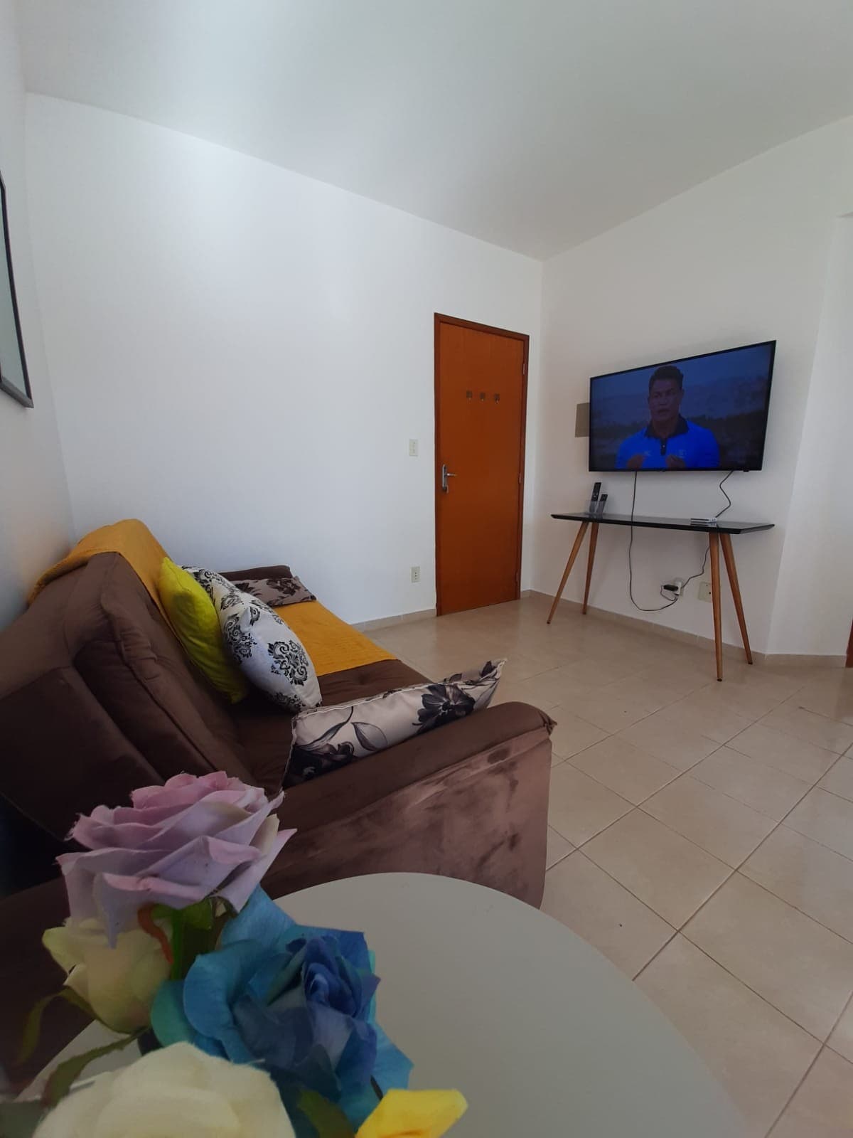 A comfortable seating area is presented with a soft brown sofa adorned with decorative pillows. A wall-mounted television is positioned opposite a simple side table, which holds a bouquet of colorful flowers. Natural light brightens the lightly colored room through a nearby door.