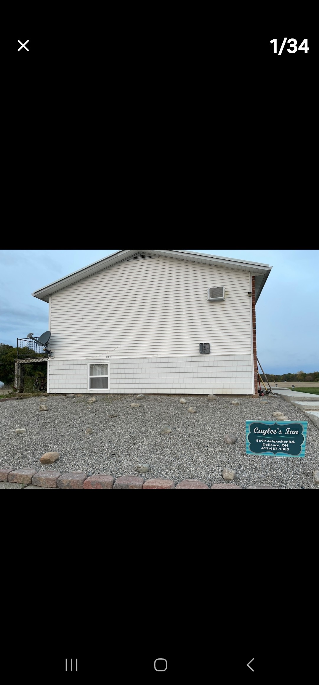 The side of the house is shown, featuring a light-colored exterior with horizontal siding. A small window and an air conditioning unit are visible. A stone gravel area surrounds the foundation, and a sign with the property's name is placed nearby.