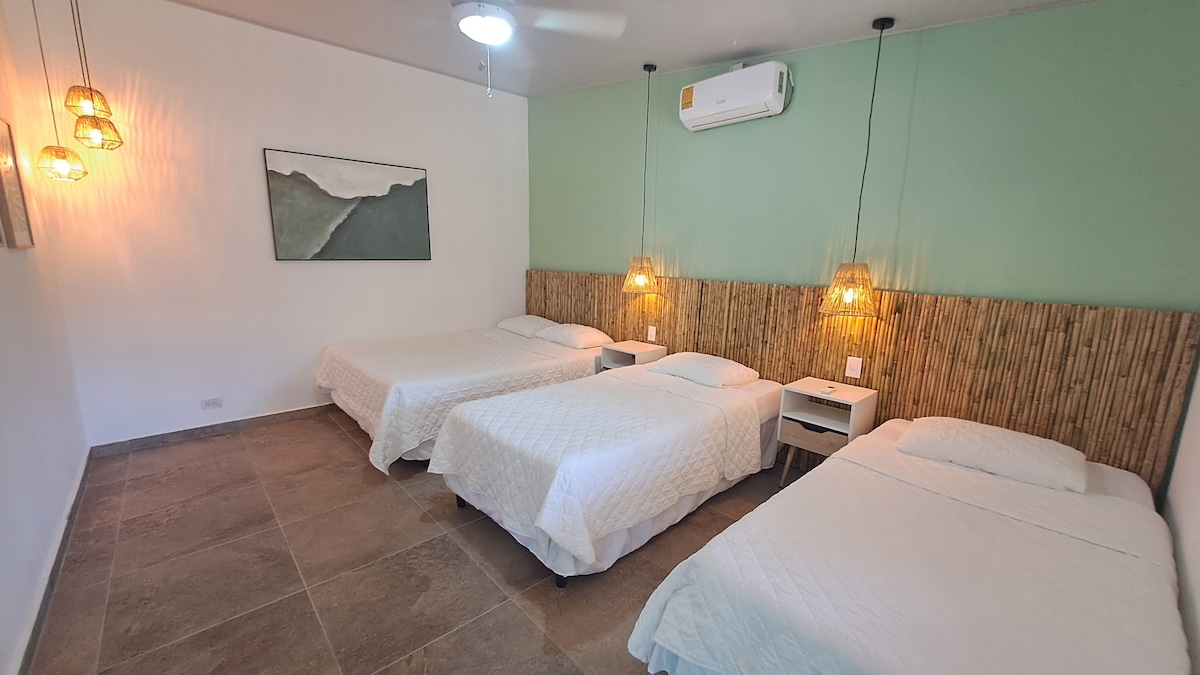 A sleeping area features three spacious beds arranged neatly. A textured bamboo headboard adds warmth to the room, complemented by simple bedside tables. Soft lighting is provided by pendant lamps, and a large artwork adorns one wall, while a ceiling fan hangs above for comfort.