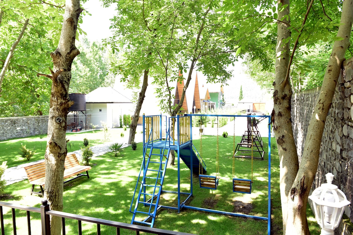 A spacious outdoor area is surrounded by lush greenery, featuring a bright blue play structure and swings for children. There are wooden benches for seating, and the scene is complemented by well-maintained grass and trees providing natural shade.