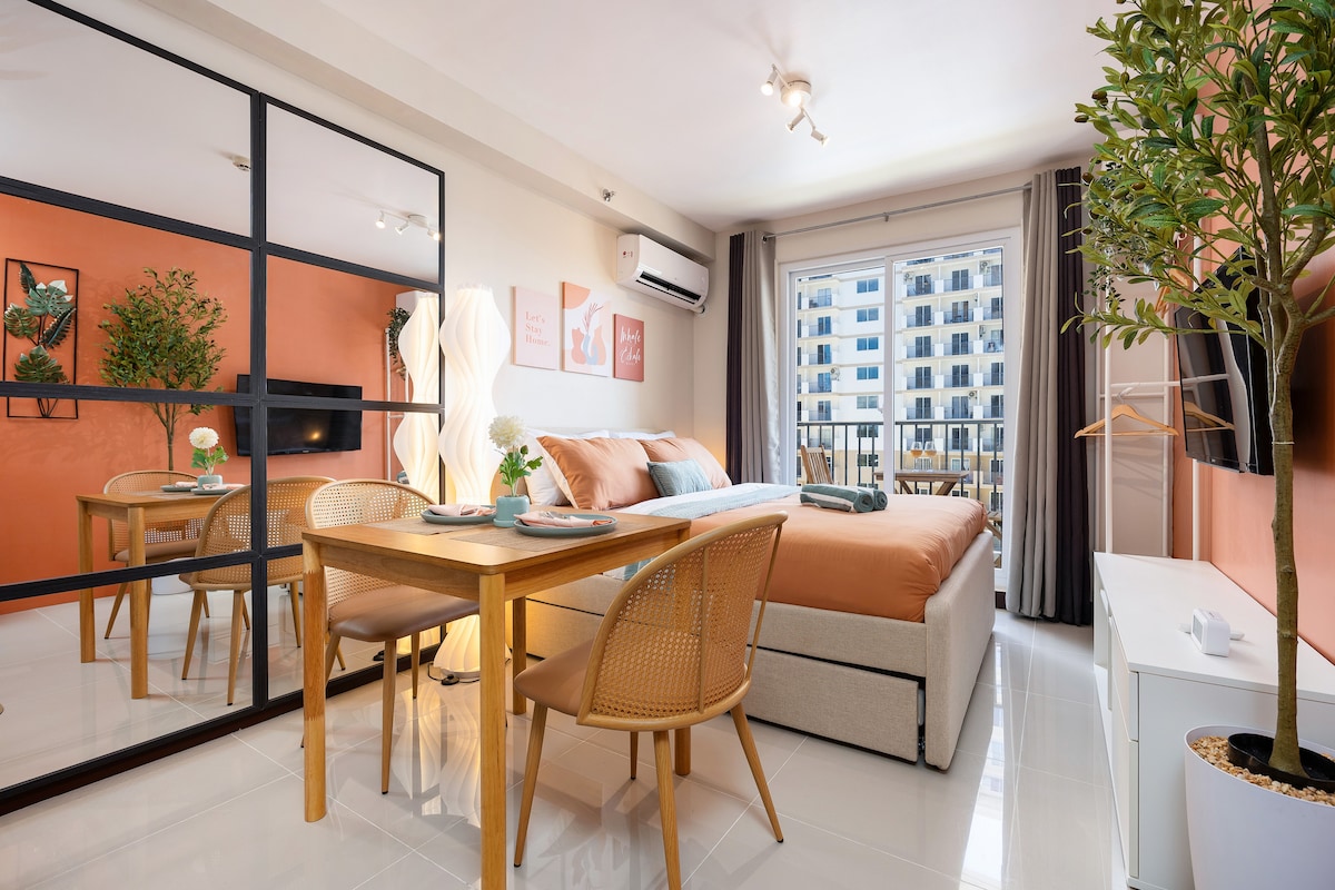 A stylish room features a spacious bed with neutral bedding, complemented by a circular dining table with rattan chairs. A large mirror creates an illusion of space, while natural light floods in through the balcony doors. Greenery adds a touch of life to the space.