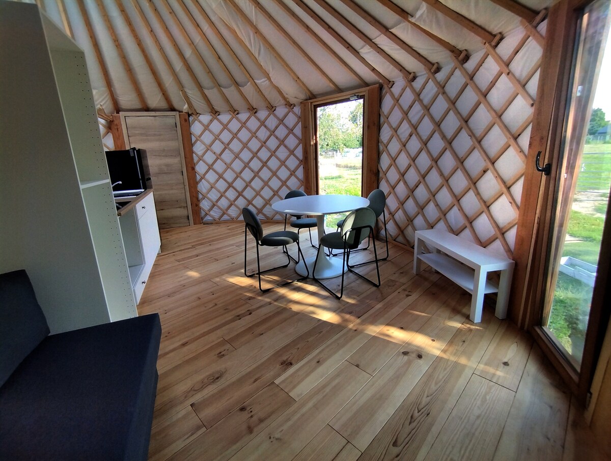 A sunny interior of a yurt features light wooden walls and flooring. A round dining table surrounded by four chairs is centrally positioned. A small white side table and a dark sofa are visible in the space. Large windows provide natural light and views of the surrounding landscape.