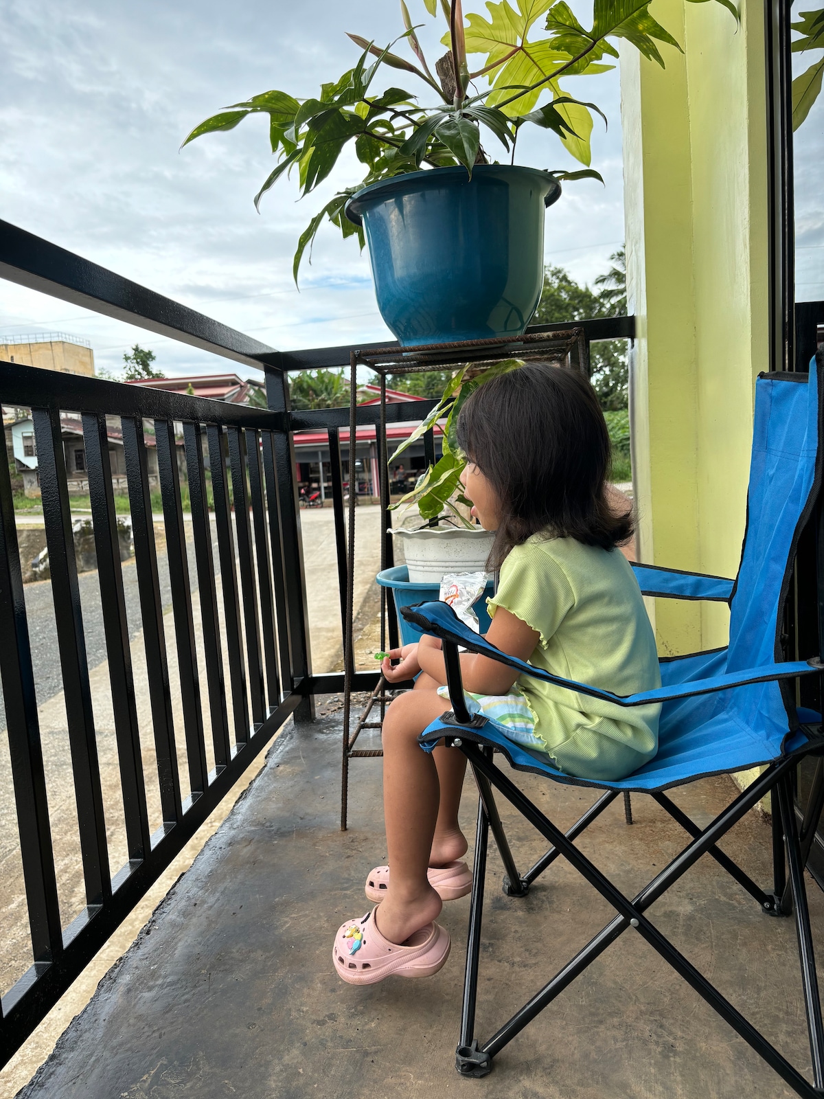 A small child sits in a blue folding chair on a balcony, observing the surroundings. A green plant is suspended in a blue pot above them. A solid black railing provides safety, while the ground below is visible, and a road is seen in the background.