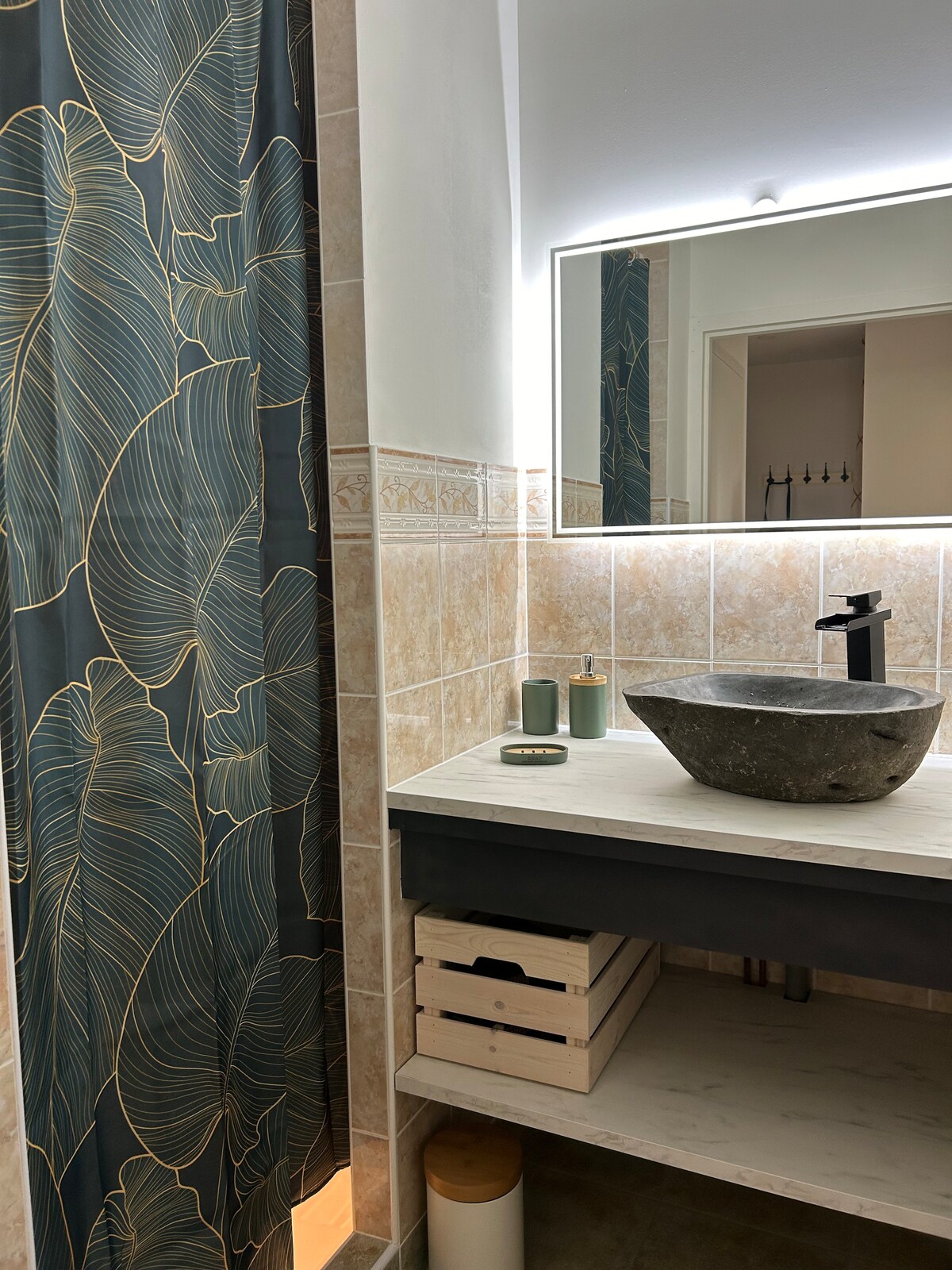 The bathroom features a modern design with a stone sink atop a light countertop. A large mirror reflects soft lighting, while a floral patterned shower curtain adds a touch of warmth. Storage is provided by a wooden crate beneath the sink.