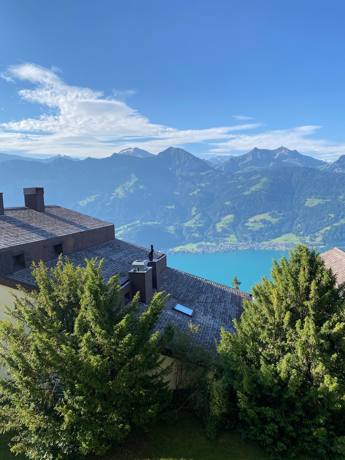 A panoramic view of the surrounding mountains and lake is captured from a vantage point. Green trees frame the scene, while rooftops of the accommodation blend into the hillside. The sky is mostly clear, breathing life into the tranquil landscape.