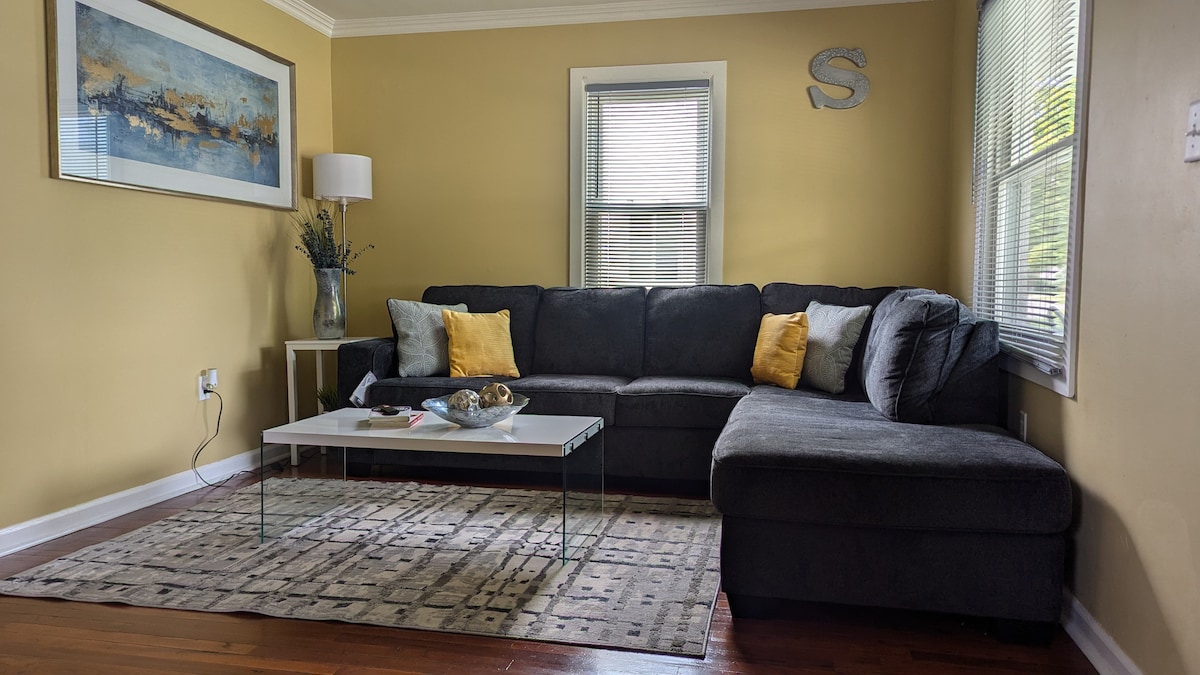A comfortable living room features a large sectional sofa adorned with yellow cushions. A glass coffee table rests on a patterned area rug. Neutral walls are complemented by a framed artwork and a vase with greenery. Natural light enters through the windows, enhancing the inviting space.