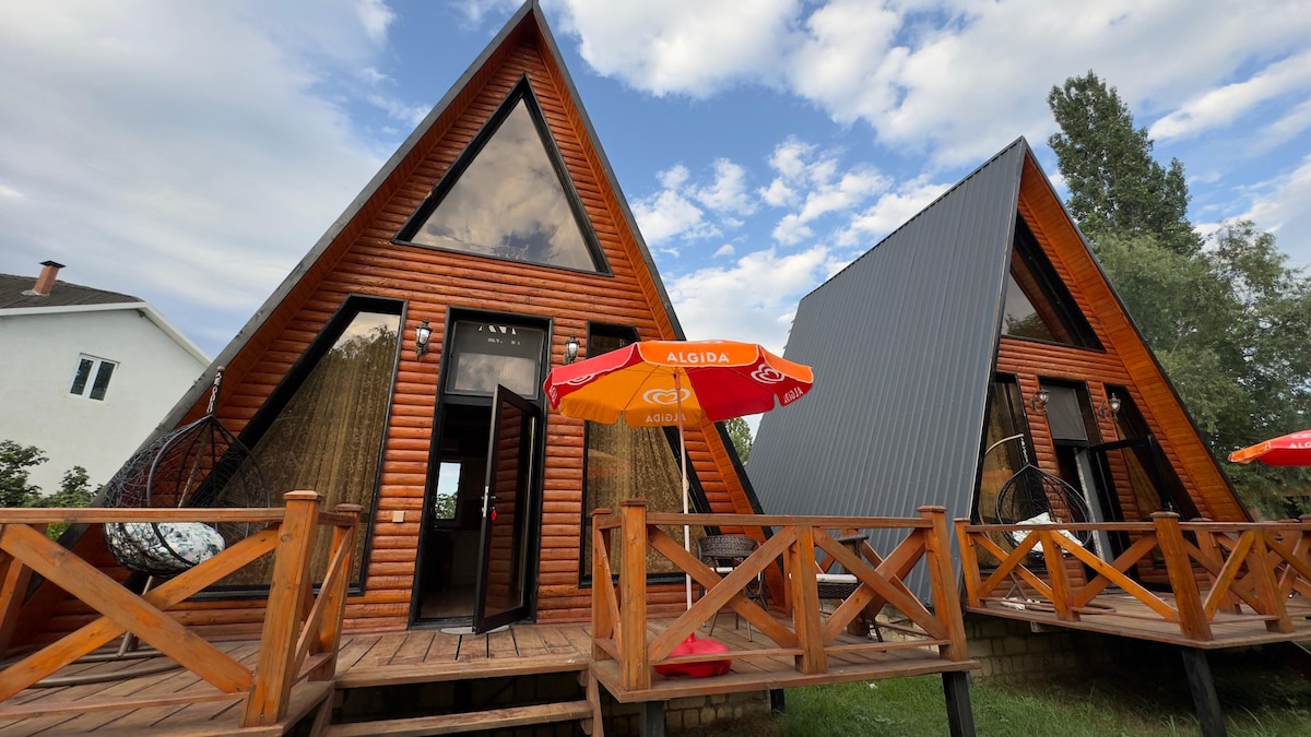 Two A-frame cabins are set among greenery, featuring large triangular windows that allow natural light to fill the interiors. Wooden decks are connected, each adorned with hanging chairs. A vibrant orange umbrella provides shade over the outdoor seating area, creating a relaxed atmosphere.