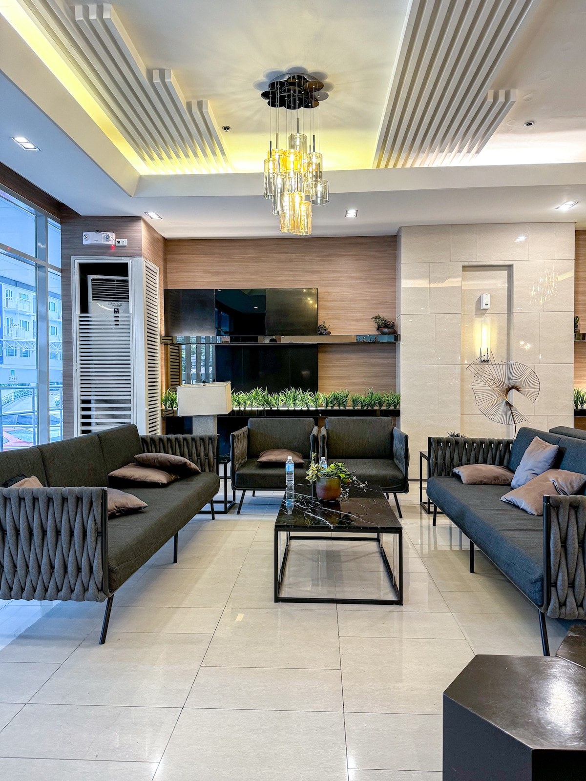 A modern living area is depicted with two cozy seating arrangements featuring dark, upholstered sofas. A sleek black coffee table sits centrally, adorned with decorative items. Soft lighting is provided by a stylish ceiling fixture, complemented by subtle greenery in the background.