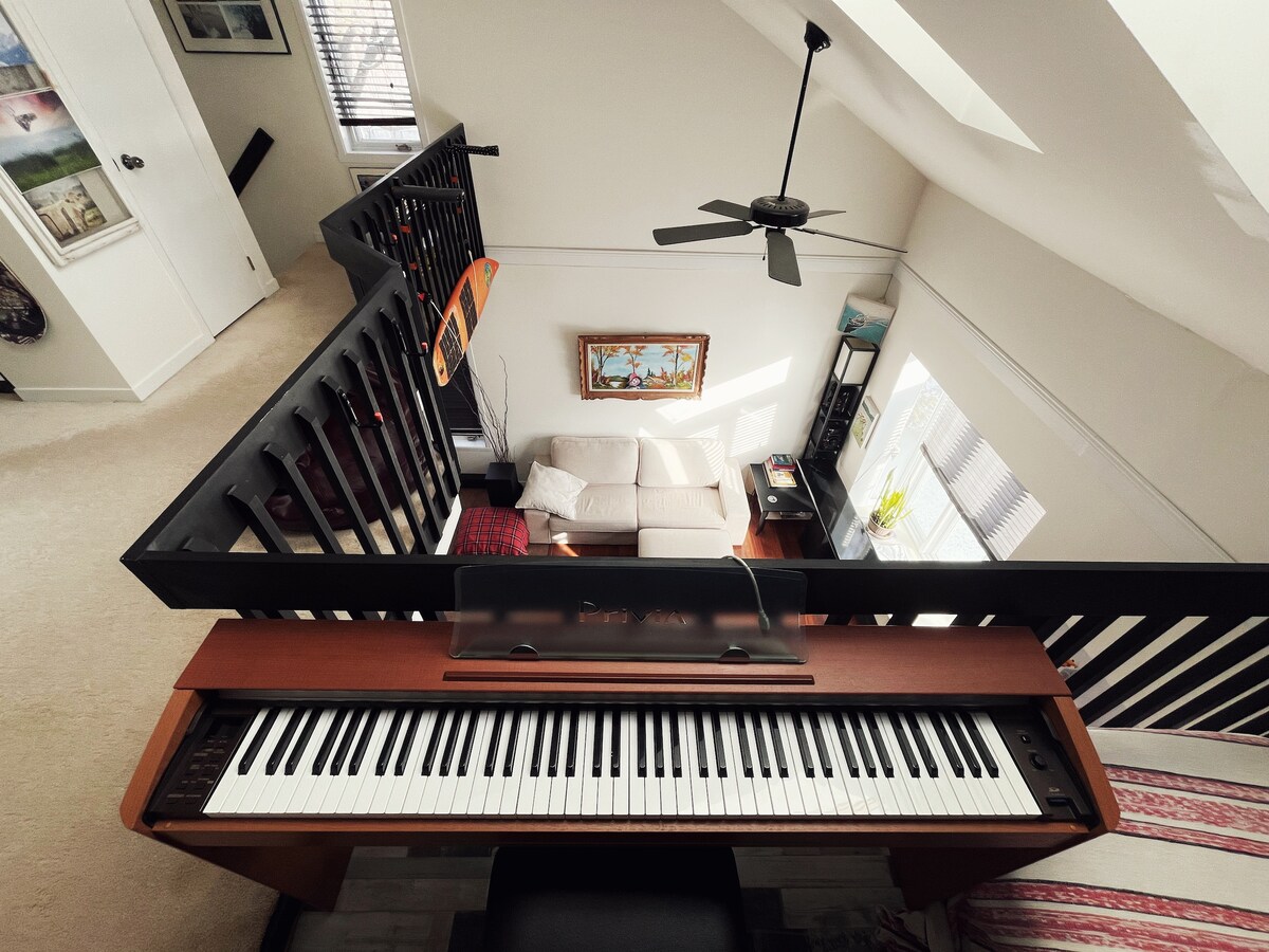 A view from above showcases a piano at the forefront overlooking a cozy living area. The space features a light-colored sofa and artwork on the wall, while soft natural light filters through a nearby window.