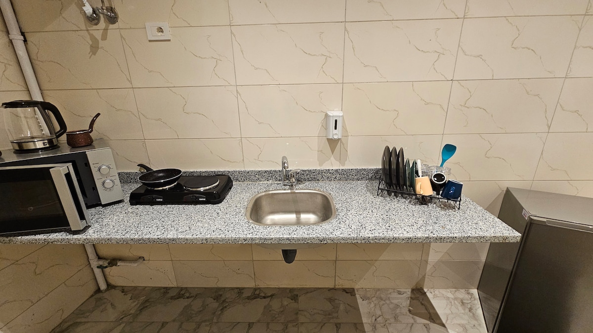 The kitchen features a granite countertop with essential appliances including a kettle, microwave, and stove. A stainless steel sink is positioned centrally, accompanied by a dish rack displaying various utensils. Light-colored tile walls create a clean and organized cooking space.