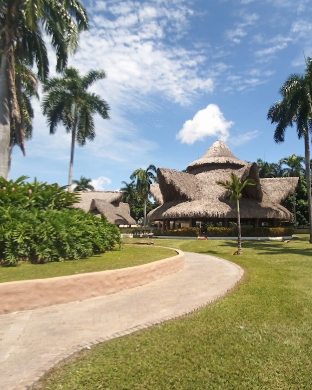 A spacious vacation property is showcased with a thatched roof and distinctive architecture, surrounded by lush palm trees and well-maintained grass. A winding pathway leads up to the structure, enhancing the natural landscape and inviting a sense of relaxation and ease.