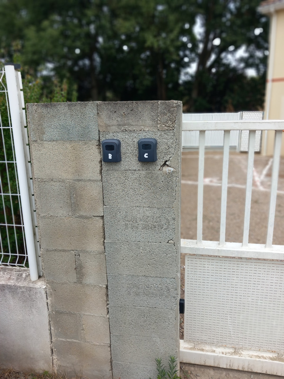 A concrete wall serves as a boundary, featuring two grey keypads mounted on it. The surrounding greenery is visible, along with a gated entrance that provides access to the property. The ground is composed of gravel, contributing to the property's outdoor environment.