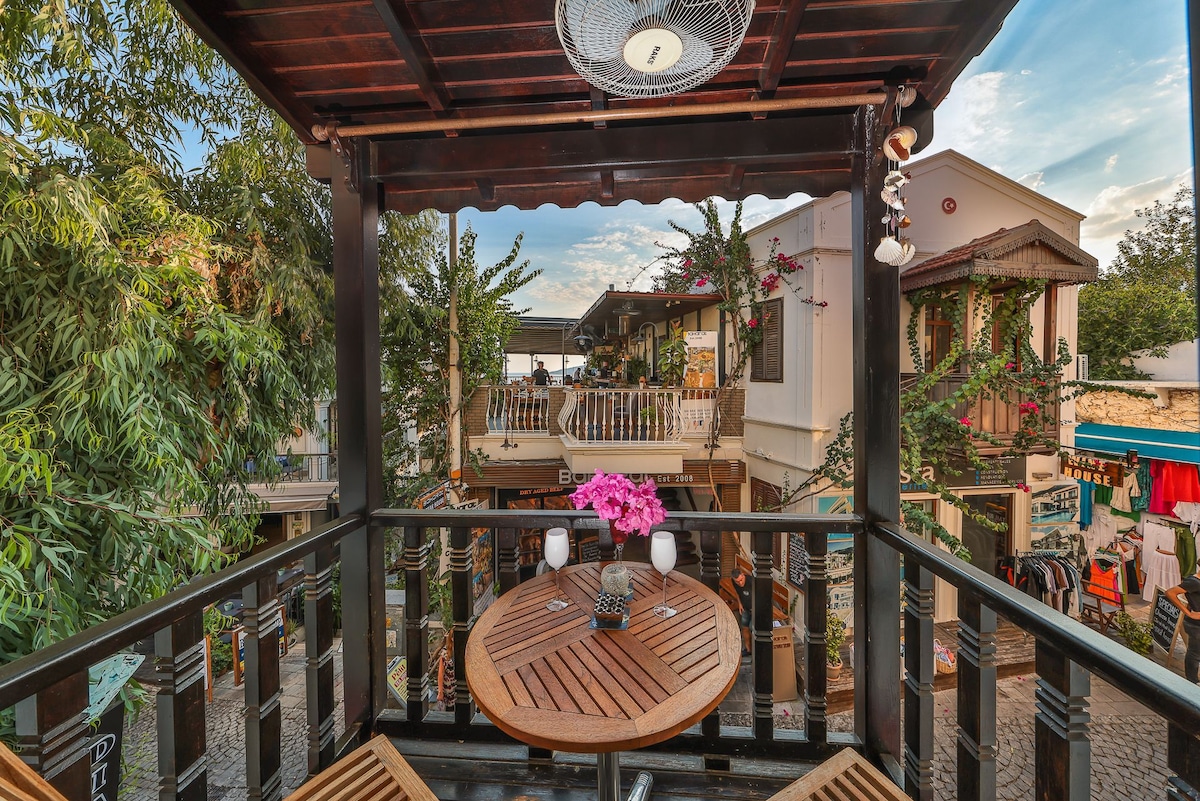 A balcony area features a round wooden table set for two, complemented by two wooden chairs. Small potted plants and pink flowers are arranged on the table, while the view reveals charming shops and greenery in the surrounding area.