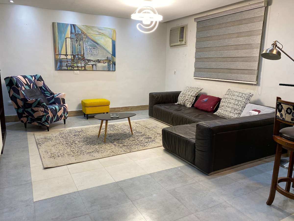 A spacious living area is highlighted by a large dark sofa and a colorful patterned armchair. A low coffee table sits in front of the sofa, centered on a soft area rug. An abstract painting adorns the wall, with natural light filtering through a window covered by horizontal blinds.