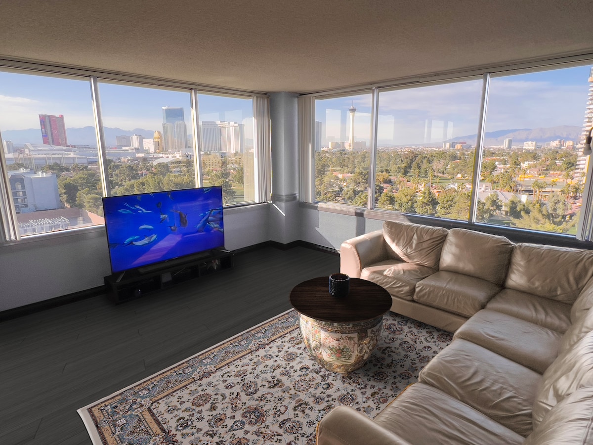 A spacious living area features a light-colored sectional couch, accompanied by a round coffee table. A large television stands on a media console, with floor-to-ceiling windows showcasing a city skyline and distant mountains, allowing ample natural light.
