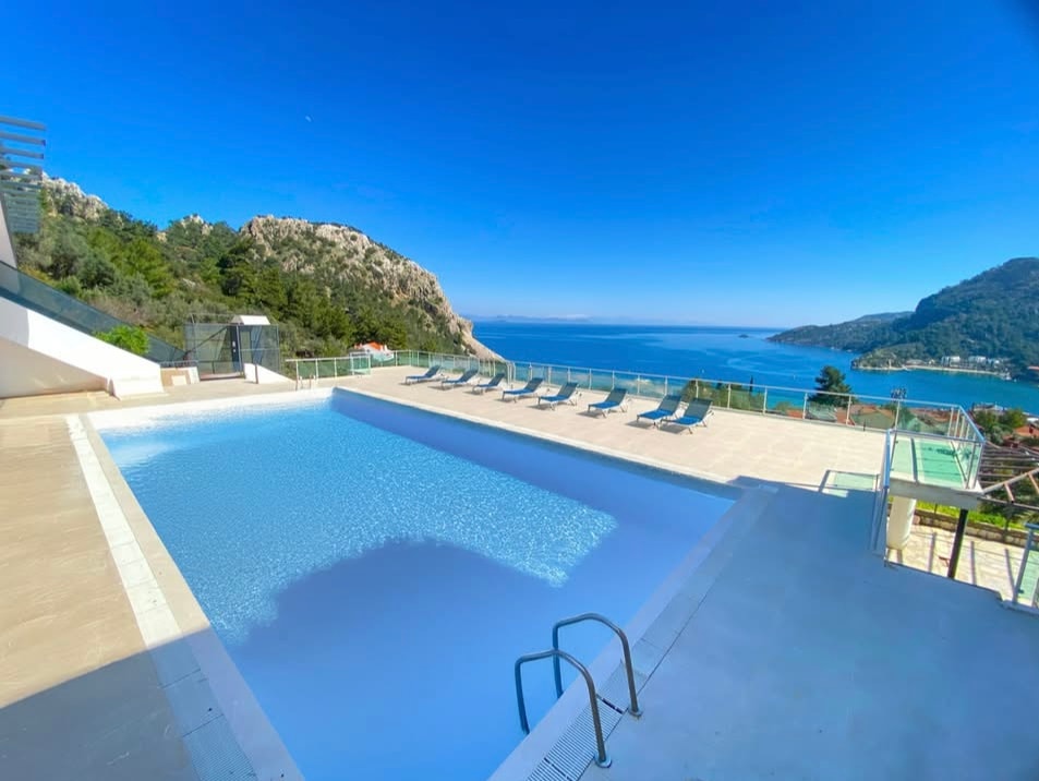An outdoor swimming pool offers expansive views of the surrounding hills and coastline. A series of lounge chairs are arranged along the pool's edge, and clear blue waters reflect the bright sky. The area is well-lit and designed for relaxation.