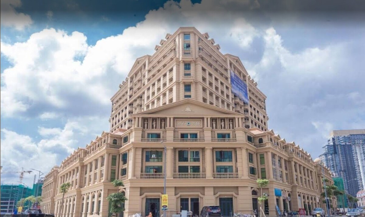 A grand building is presented with classic architectural features, including multiple stories and detailed balconies. Large windows allow for natural light, while palm trees are positioned along the entrance, contributing to a welcoming exterior against a backdrop of clouds.