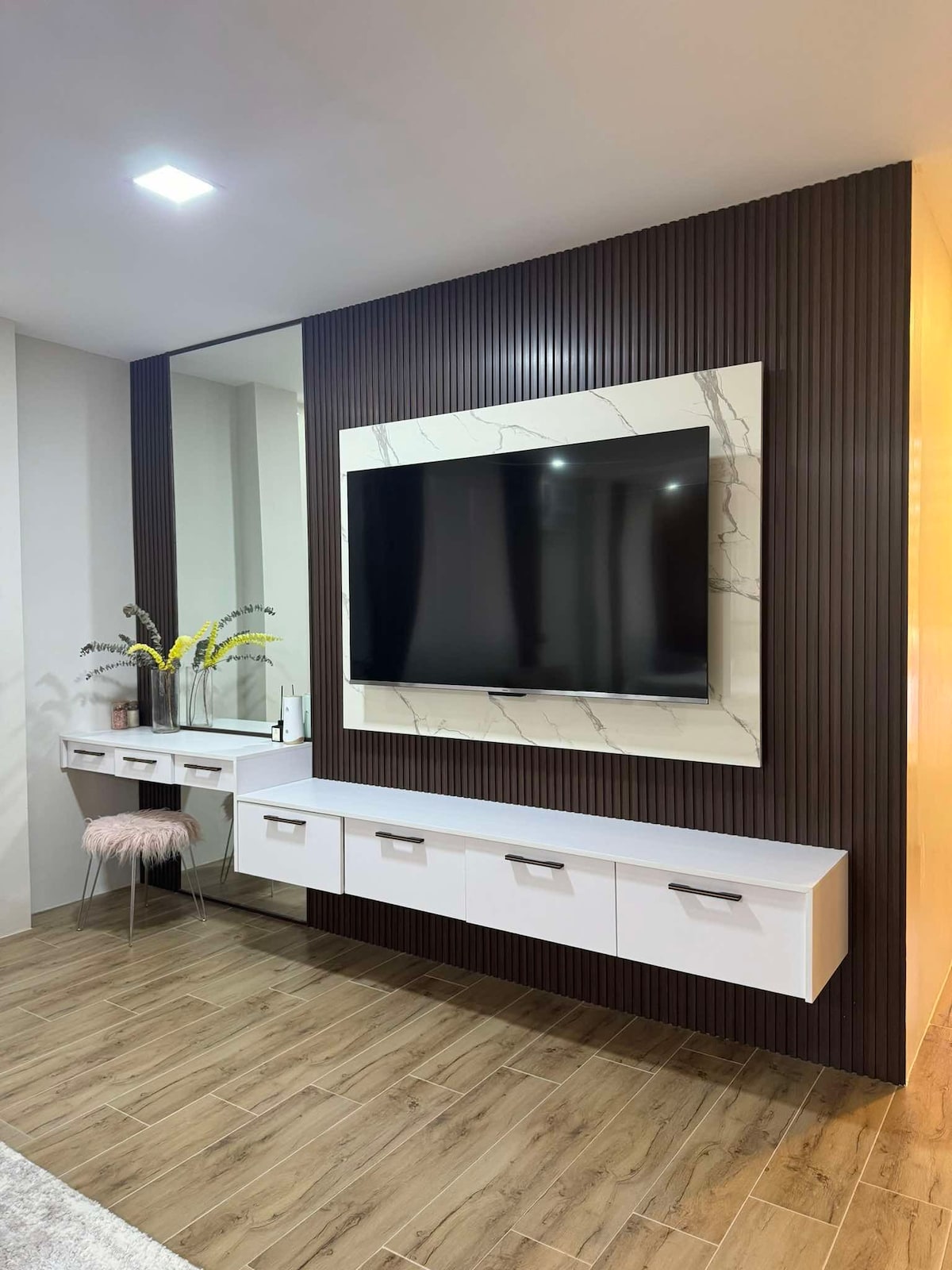 A wall-mounted television is framed by a modern white and marble panel. Below, multiple drawers provide storage space, while a decorative mirror reflects light. A small, plush ottoman adds comfort to the area, against a backdrop of rich, dark paneling.