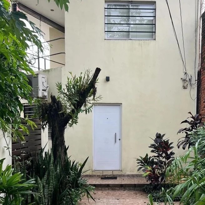 A simple white door stands at the center of a light-colored wall. Lush green plants surround the entrance, providing a natural, inviting touch. A large tree, partially visible on the left, adds to the greenery of the area, which appears serene and well-maintained.