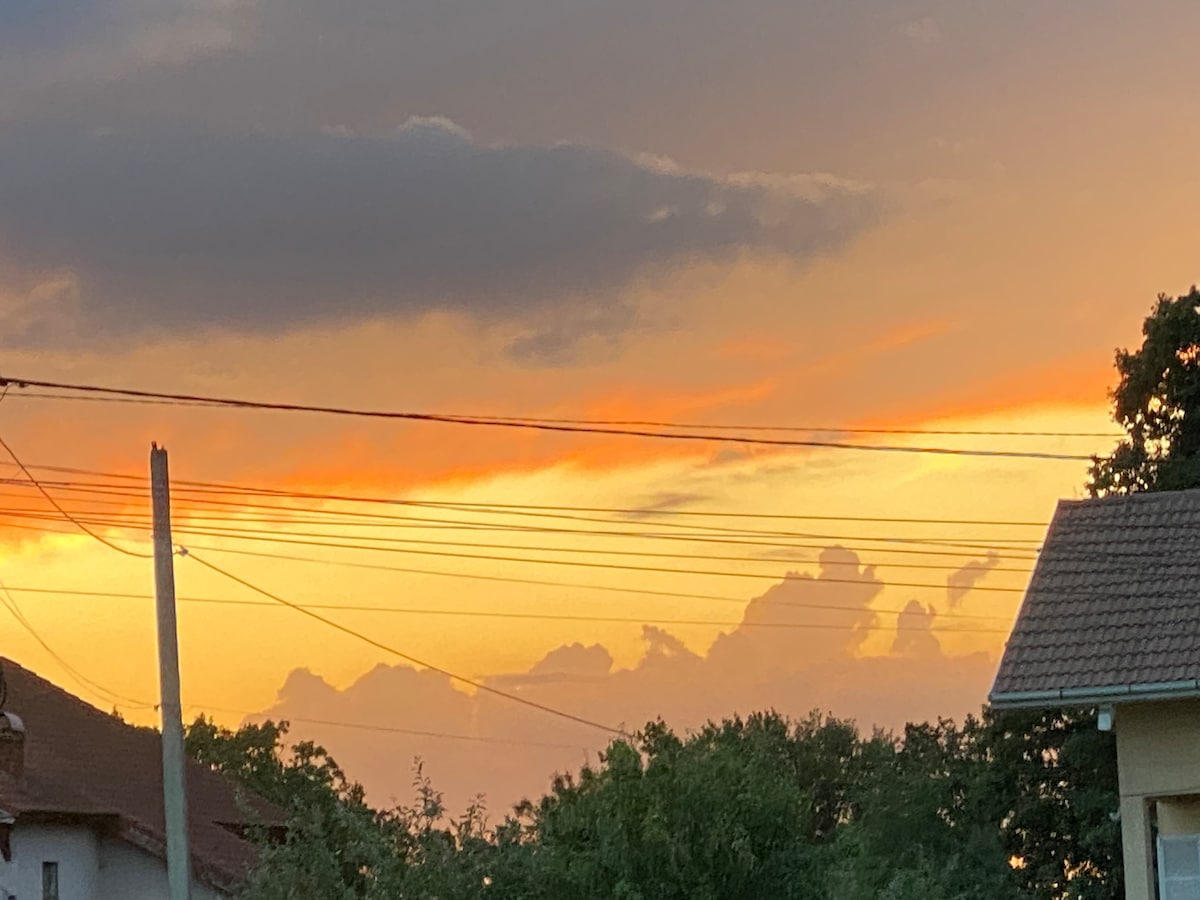A vibrant sunset is captured, showcasing hues of orange and yellow blending with soft clouds. A silhouette of houses is visible in the foreground, with power lines intersecting the sky, enhancing the scenic view.